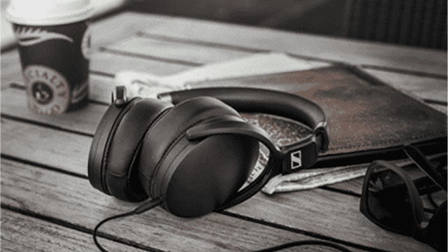 Zwarte hoofdtelefoon naast een bruine portemonnee, een zonnebril en een papieren beker op een houten tafel.