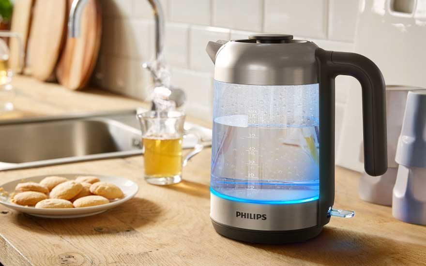 Glazen Philips-waterkoker met blauwe verlichting kookt water op een houten aanrecht, naast een kop thee en een bord met ronde koekjes.