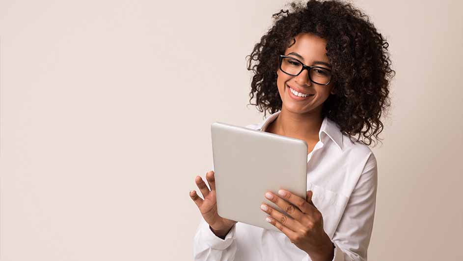 Een vrouw met een witte blouse en bril lacht terwijl ze een tablet vasthoudt, voor een beige achtergrond.