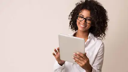 Een vrouw met een witte blouse en bril lacht terwijl ze een tablet vasthoudt, voor een beige achtergrond.