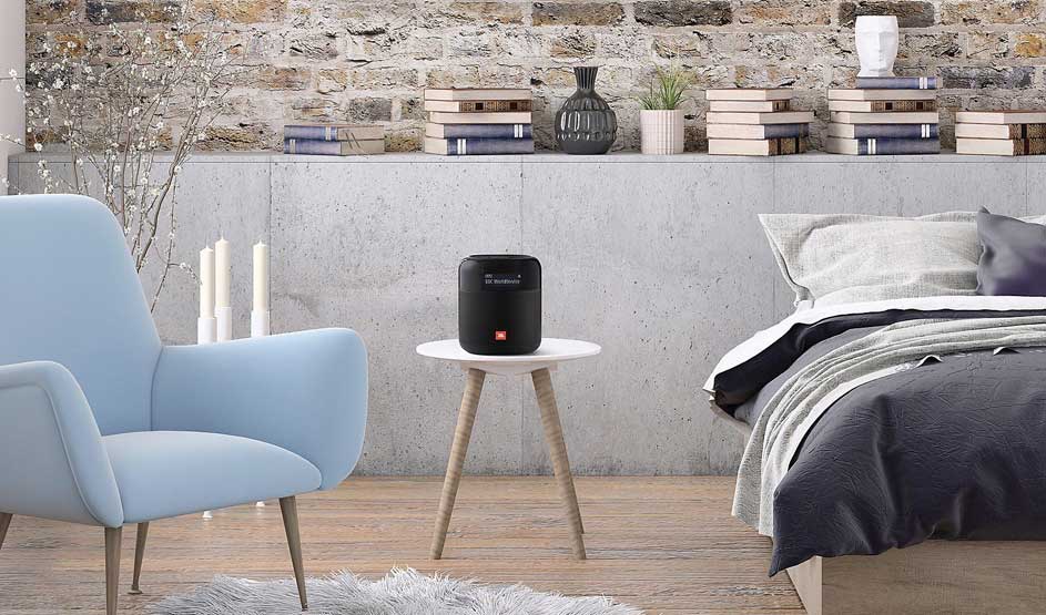 Op een wit tafeltje naast een bed staat een zwarte JBL speaker in een moderne slaapkamer met boeken, planten en een blauwe fauteuil.
