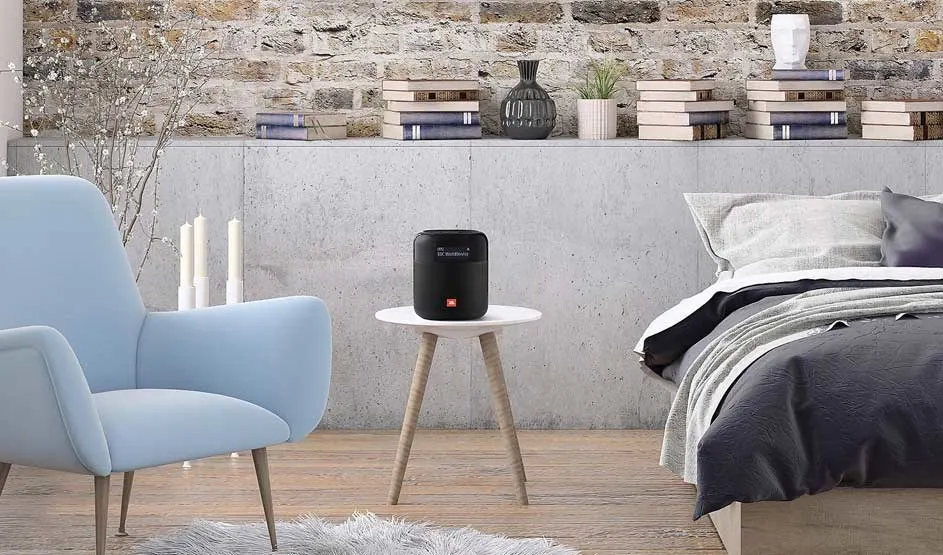 Op een wit tafeltje naast een bed staat een zwarte JBL speaker in een moderne slaapkamer met boeken, planten en een blauwe fauteuil.