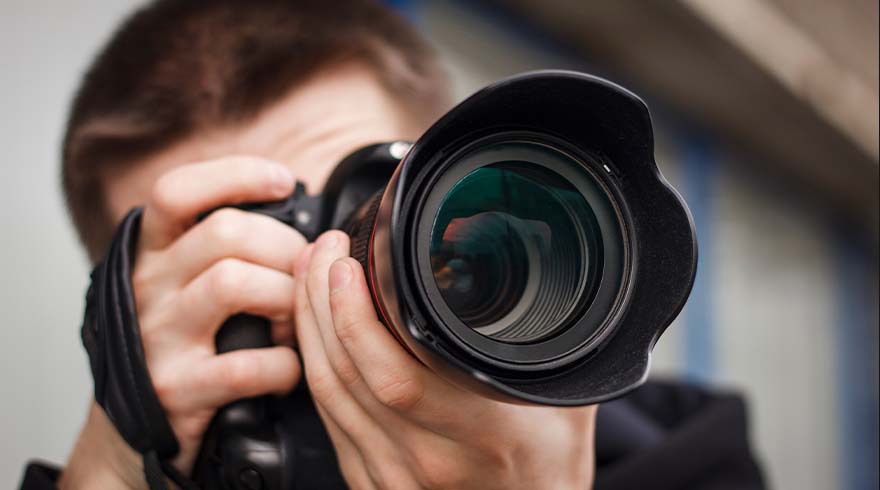 Een persoon houdt een zwarte camera met een grote lens vast, gericht naar voren. De achtergrond is lichtgrijs.