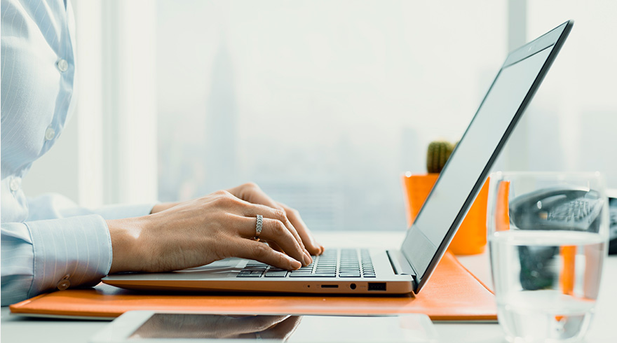 Vrouw typt op een laptop op een oranje onderlegger. Er staat een glas water en een plant in een oranje pot op de achtergrond.