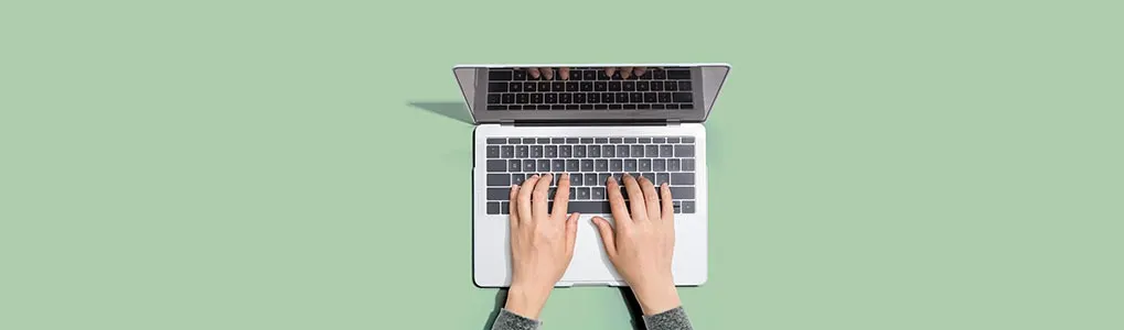 Overhead shot of hands typing on a silver laptop against a light green background.