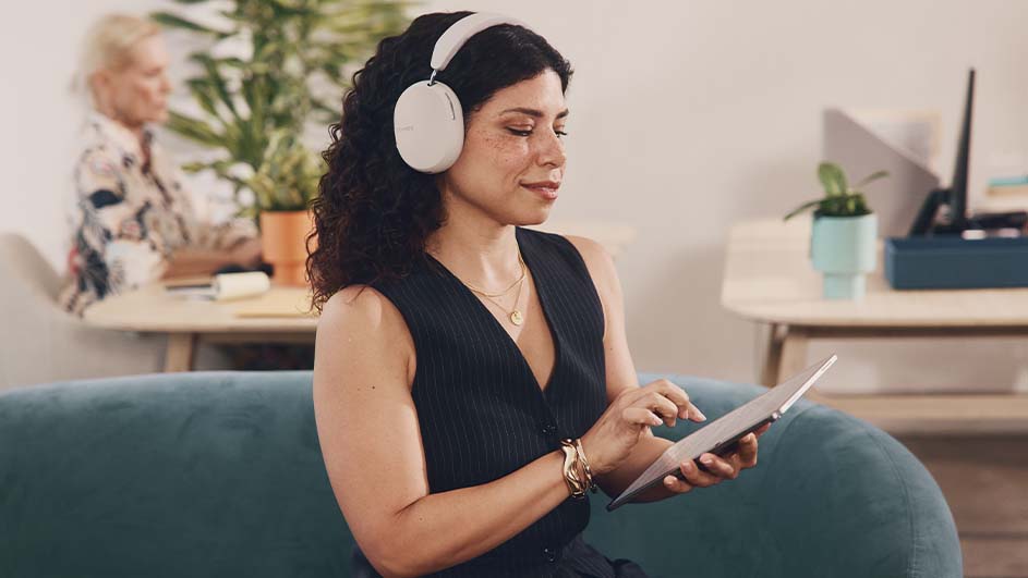 Vrouw met hoofdtelefoon gebruikt een tablet op een groene bank. Achter haar staat een bureau met planten en een persoon.