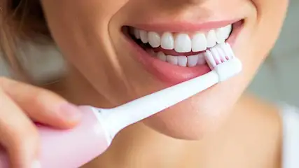 Close-up of a smiling woman brushing her teeth with a pink electric toothbrush in a bright, clean bathroom setting.