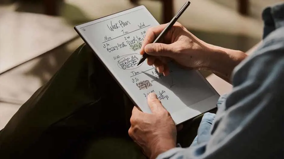 Iemand schrijft met een pen in een weekplanner op een tablet, zittend op een bank.