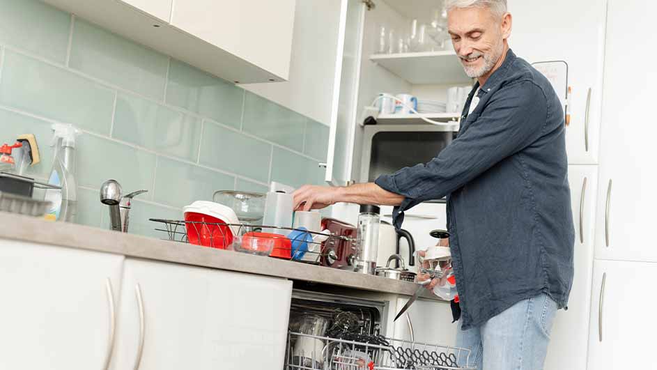 Een man laadt een vaatwasser in een keuken met witte kasten en blauwe tegels. Op het aanrecht staat een afdruiprek.