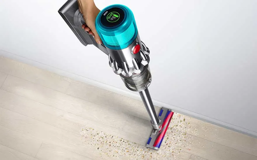 Bovenaanzicht van een steelstofzuiger die kruimels van een lichte houten vloer opzuigt, met gekleurde borstelkop en transparante opvangbeker.