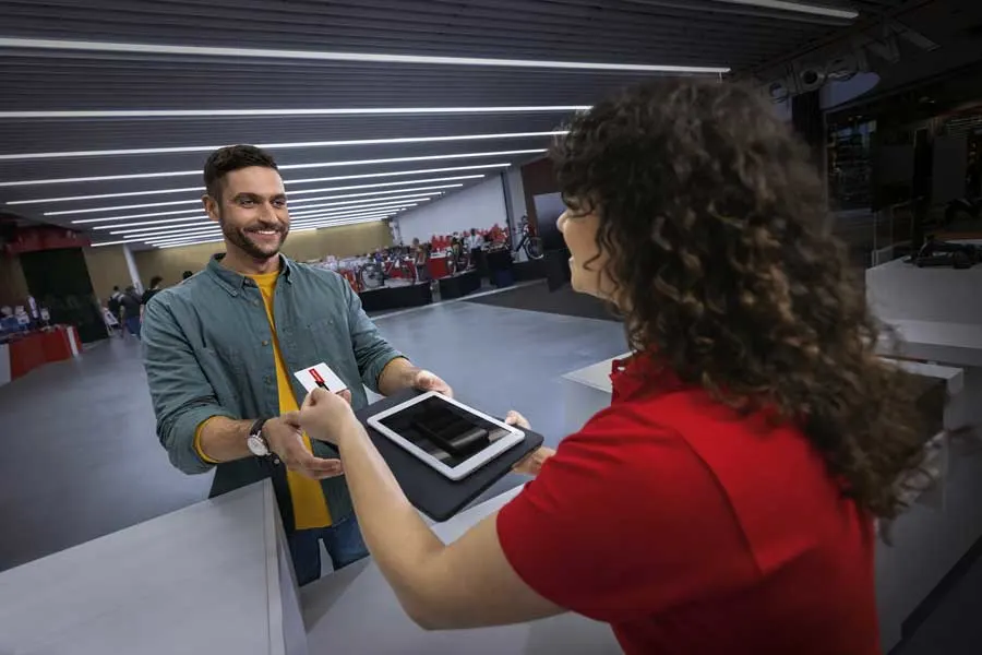 Een medewerker in rood shirt overhandigt een tablet en kaart aan een klant bij een balie in een moderne winkel met veel licht en displays.