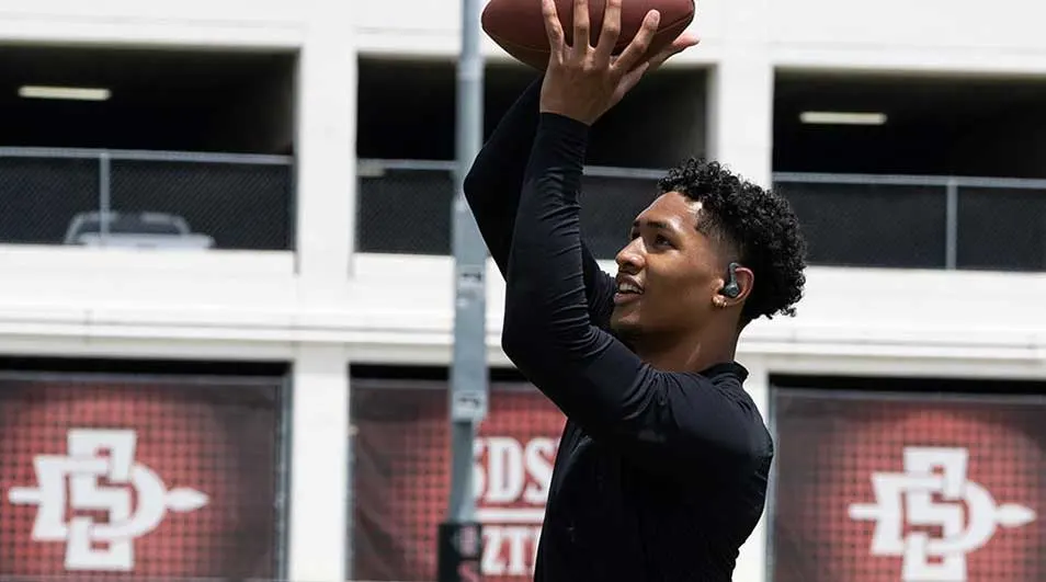 Een jonge man speelt American football buiten, reikt hoog om de bal te vangen terwijl hij draadloze oortjes draagt en gefocust naar voren kijkt.