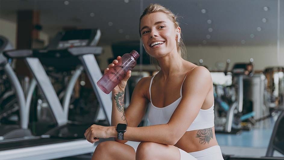 Vrouw met witte sportkleding en smartwatch houdt roze waterfles vast in een fitnessruimte met loopbanden op de achtergrond.