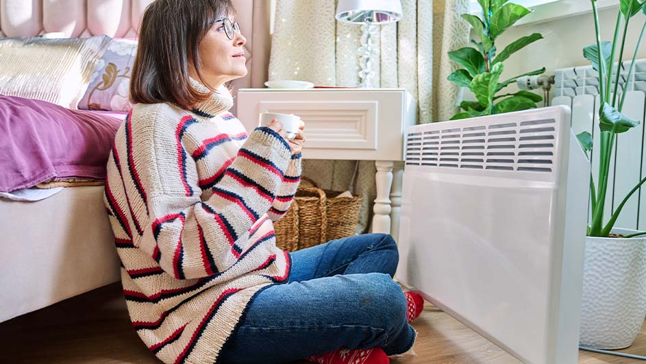 Een vrouw zit op de vloer met een kopje, naast een witte radiator en een plant. Een bed en een nachtkastje staan achter haar.