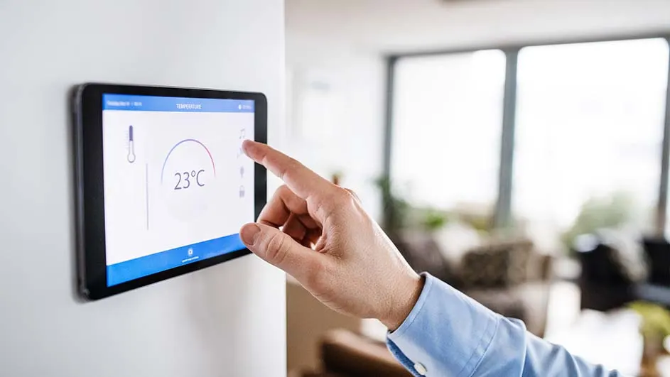 Een hand bedient een zwart tablet aan een witte muur, waarop 23°C en een thermometer te zien zijn. Achtergrond: raam en woonkamer.