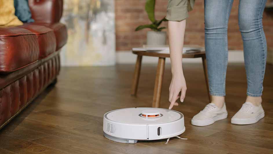 Een witte robotstofzuiger staat op een houten vloer, een hand wijst ernaar, met een bank en een tafel op de achtergrond.