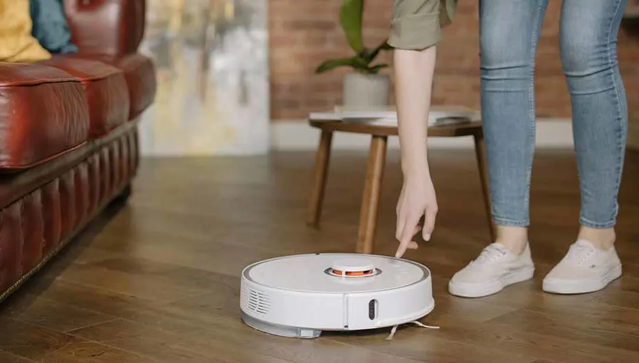 Een witte robotstofzuiger staat op een houten vloer, een hand wijst ernaar, met een bank en een tafel op de achtergrond.