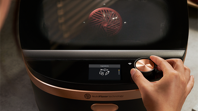 Een hand draait aan een koperen knop op een zwarte NutriFlavor-oven. Op het scherm staan pictogrammen voor vis, kip en maïs.
