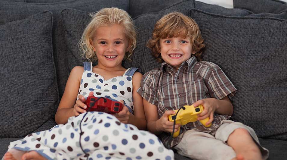 Twee lachende kinderen zitten op een grijze bank met rode en gele controllers in hun handen.