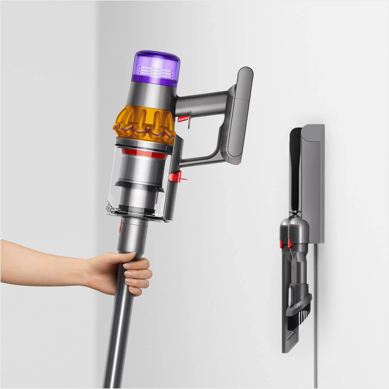 Een hand houdt een grijze Dyson stofzuiger met paarse, gouden en rode details. Een grijze wandhouder hangt aan een witte muur.
