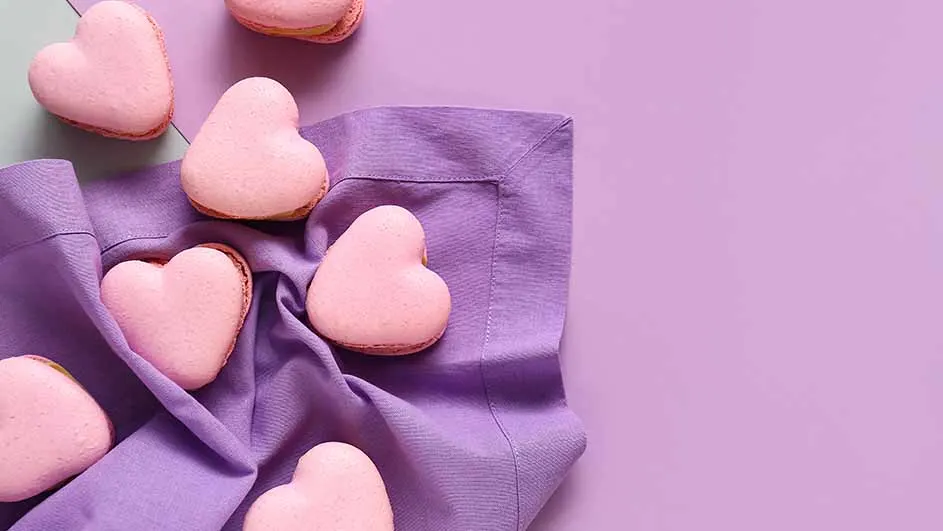 Roze hartvormige macarons op een paarse stoffen servet, tegen een paarse achtergrond.