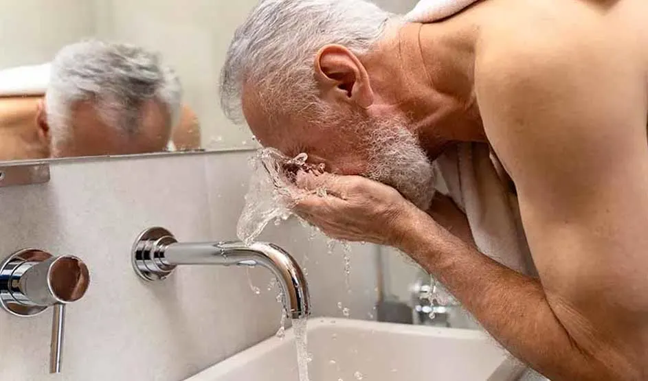 An older man bends over the sink and splashes water on his face; his reflection is visible in the bathroom mirror.
