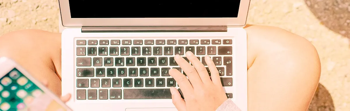 A person uses a laptop with a black keyboard, while holding a smartphone on a light brown surface.