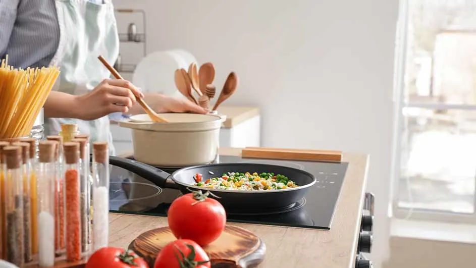 Een persoon roert in een pan en een pot op een fornuis. Er staan tomaten, pasta en kruiden op het aanrecht.