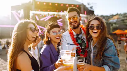 Vier lachende mensen met zonnebrillen houden bier vast. Een podium is op de achtergrond te zien.