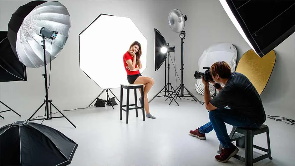 Een fotostudio met een model in een rode top op een kruk, gefotografeerd door iemand met een camera, omringd door lampen.