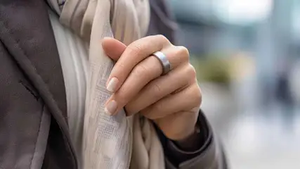 Een hand met een zilveren ring houdt een gestreepte sjaal vast, met een bruin jasje op de achtergrond.