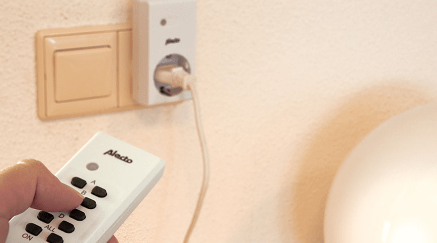 Hand bedient witte Alecto-afstandsbediening bij stopcontact en lichtbeige schakelaar aan een muur met wit snoer.