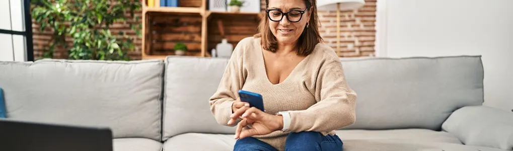 A woman wearing glasses sits on a couch, using a blue phone. Bookshelves and green plants are visible in the background.