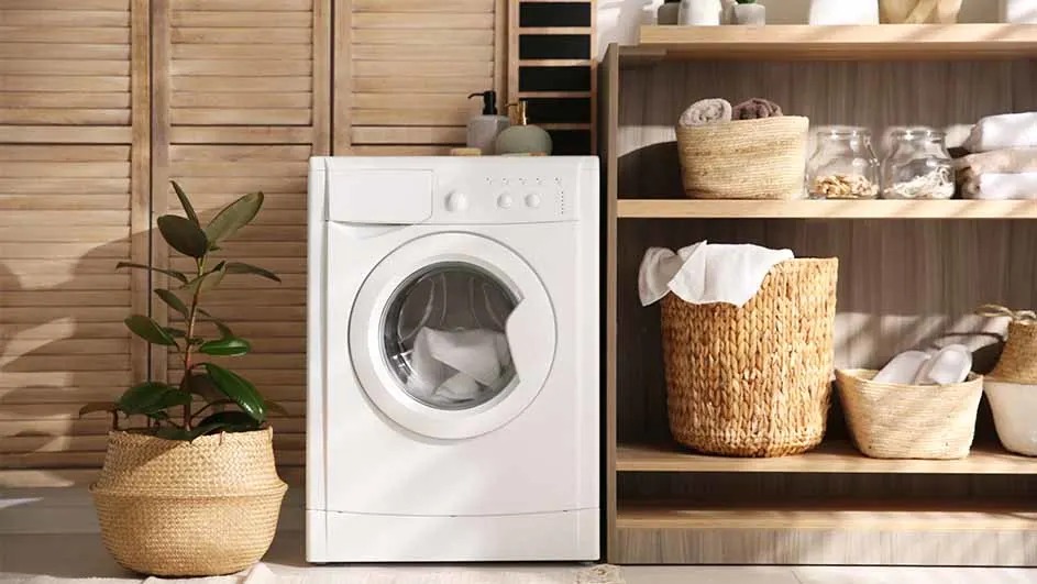 Een witte wasmachine staat naast een houten kast met manden en potten. Naast de wasmachine staat een plant in een mand.