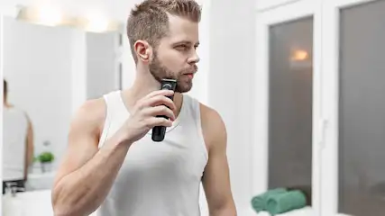Een man in een wit hemd trimt zijn baard in een badkamer met een zwart apparaat.