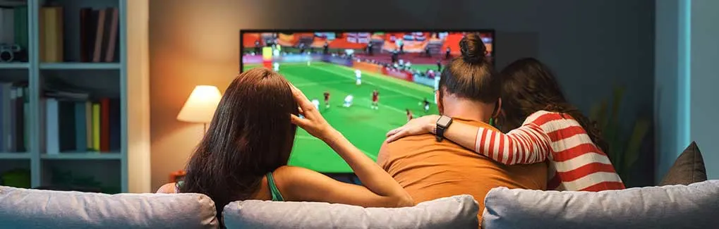 Drie mensen zitten samen op de bank en kijken knus naar een voetbalwedstrijd op tv in een warm verlichte woonkamer.