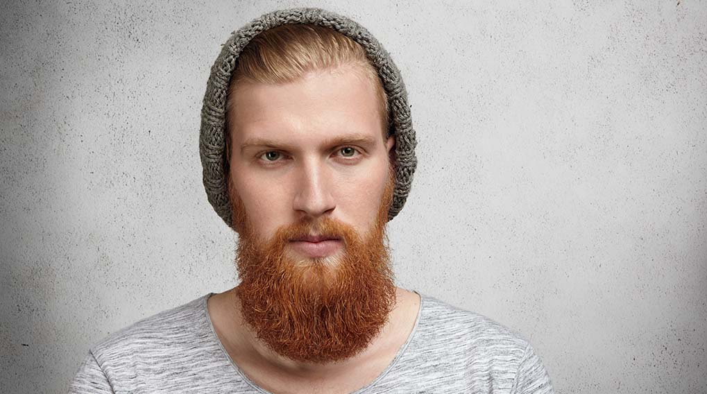 Een man met een grijze muts en een roodbruine baard staat voor een witte muur, gekleed in een grijs gestreept shirt.