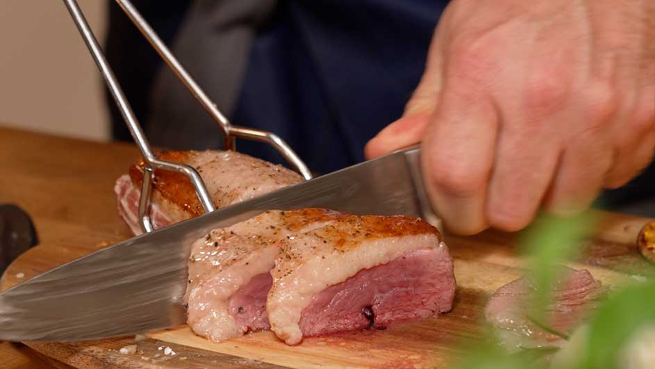 Close‑up van een hand die een stuk rosé gebraden vlees in plakken snijdt op een houten snijplank.