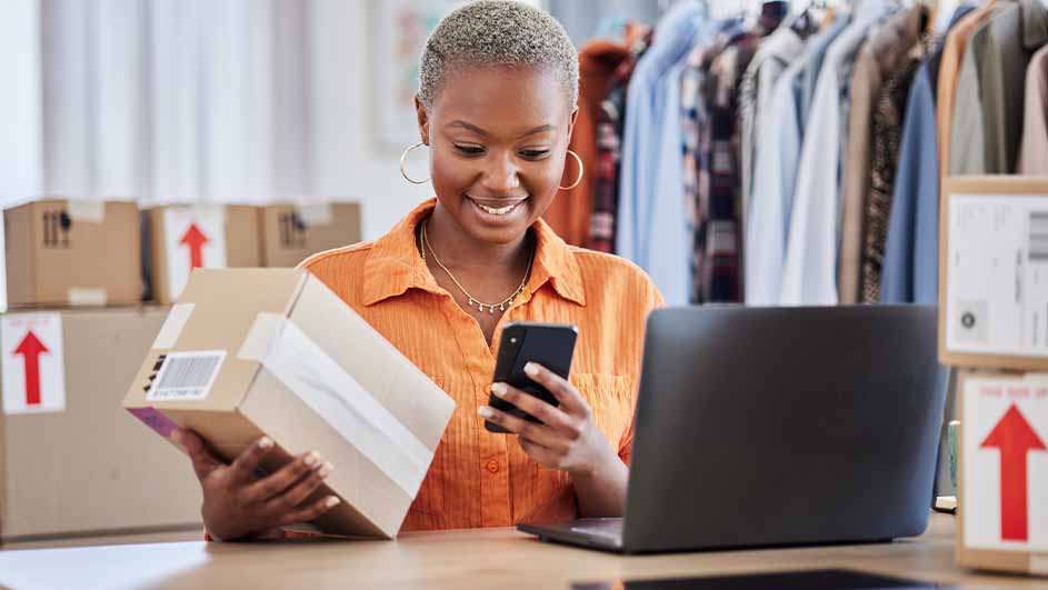 Vrouw met telefoon en pakket, laptop op tafel. Achtergrond: kleding en dozen met rode pijlen.