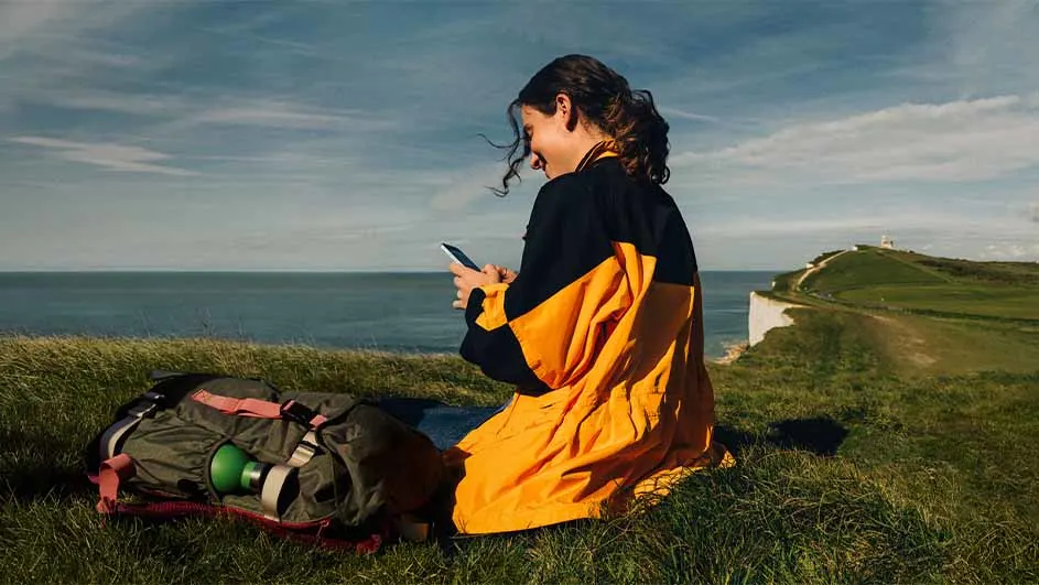 Een vrouw in een geel-zwarte jas zit met een smartphone op een grasheuvel aan zee, naast een rugzak, met kliffen en een wandelpad op de achtergrond.