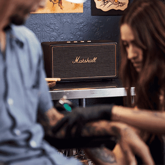 Een Marshall-luidspreker staat op een zilveren plank tegen een blauwe, patroonbehangen muur in een tattoo-studio, met een vrouw die tatoeëert.