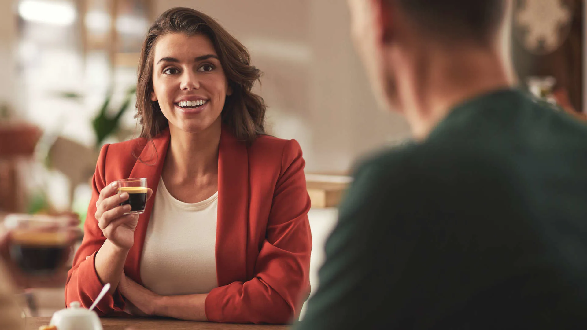 Vrouw met rode blazer houdt een glas koffie vast aan een tafel. Een man zit tegenover haar.