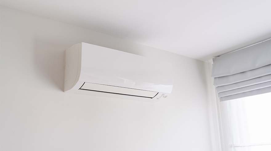 Witte airconditioner aan een witte muur, met een lichtgrijze plisségordijn rechtsboven in de hoek.