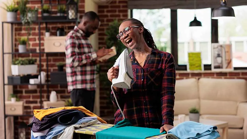 Vrouw lacht met strijkijzer in de hand bij strijkplank met kleding. Man leest boek op achtergrond. Bakstenen muur en planten.