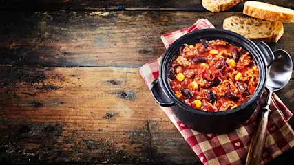 Een zwarte pan met chili staat op een rood-wit geruite doek, naast een lepel en brood op een houten tafel.