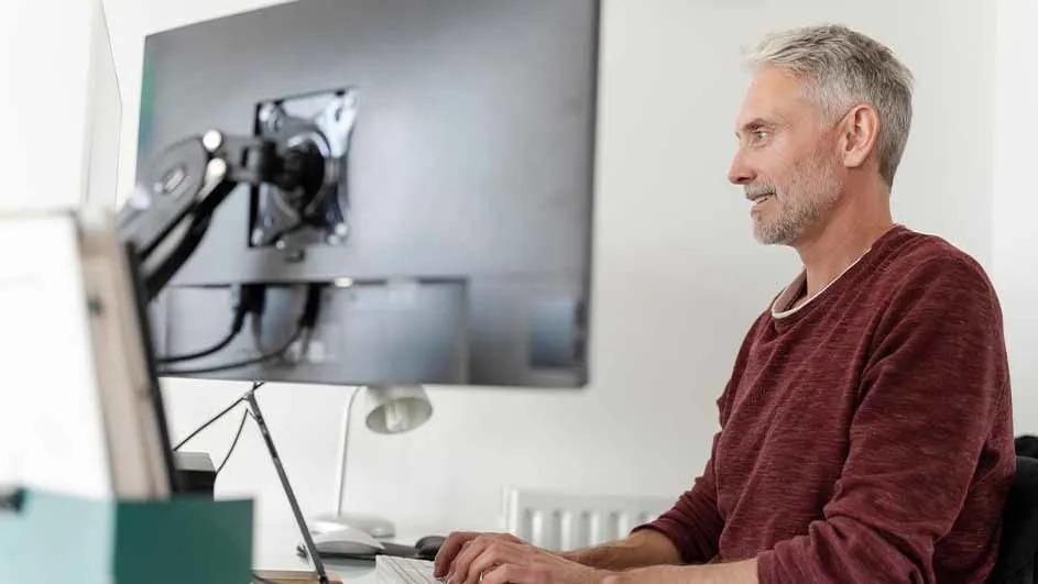 Grey‑haired man smiling while working at a large monitor on an arm, seated at a bright, modern home office desk.