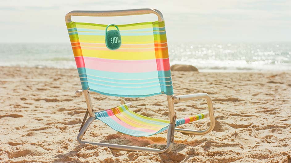 Een strandstoel met regenboogkleuren en een JBL-luidspreker staat op een zandstrand met de oceaan op de achtergrond.