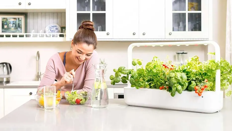 A modern kitchen featuring a smart garden full of fresh herbs and vegetables. A person prepares a salad using ingredients from the garden.