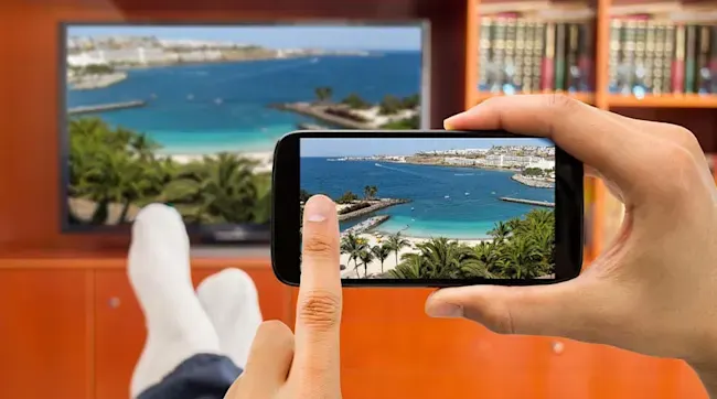 Een hand houdt een telefoon vast met een strandfoto, terwijl een tv en boekenkast op de achtergrond te zien zijn.