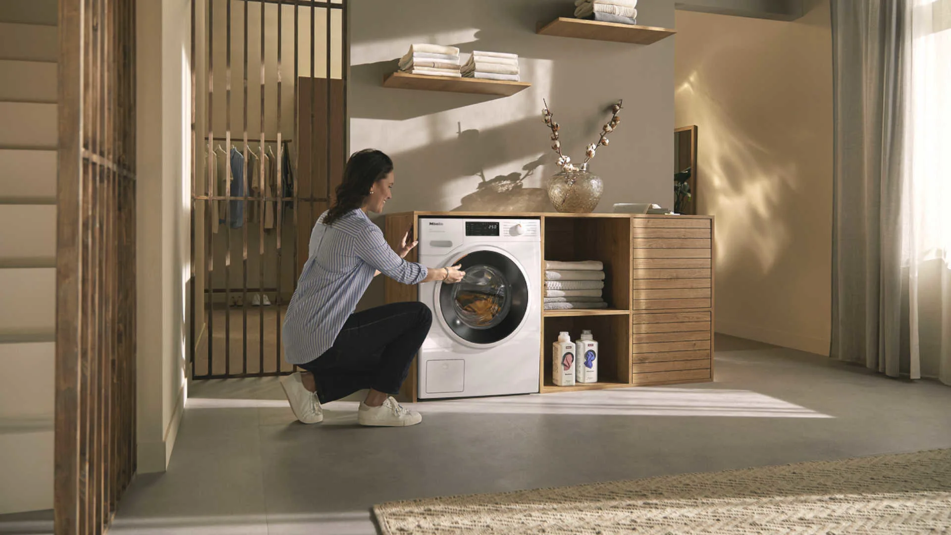 Een vrouw hurkt voor een witte wasmachine in een wasruimte met houten rekken en accessoires.